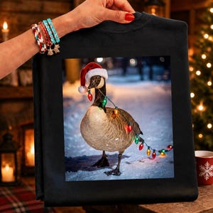 May include: A black sweatshirt featuring a graphic print of a Canada goose wearing a red and white Santa hat. The goose is wrapped in a string of colorful Christmas lights and is standing on a snowy surface. The design is centered on the fabric.