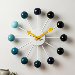May include: A modern wall clock with a white face and white spokes, each tipped with a colorful sphere. The spheres are in shades of blue and black. The clock hands are yellow.