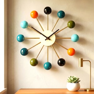 May include: A wall clock featuring a central cream-colored circular face with two dark brown hands. Twelve thin gold-toned metal spokes radiate outward, each tipped with a smooth, glossy sphere in various colors including orange, teal, olive, and black.