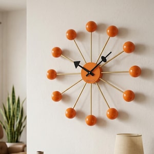 May include: A modern wall clock with a unique design. The clock features a round orange face with black hands and gold-colored spokes. Each spoke ends with a round orange ball. The clock is mounted on a white wall.