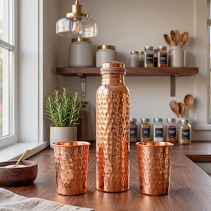 May include: A copper water bottle and two matching cups with a hammered texture. The bottle has a screw-on lid. The items are displayed on a wooden countertop in a kitchen setting, with a plant and spices in the background.