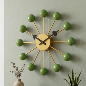 May include: A mid-century modern wall clock with a gold-colored face and arms. The clock features green spherical accents and black hands. The clock is mounted on a light green wall. The clock's diameter is approximately 18 inches.