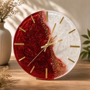 Peut inclure: Une horloge murale ronde au design divisé en résine rouge profond et blanc nacré. La surface présente une texture tourbillonnante avec des accents de feuilles d'or le long de la séparation centrale. Elle inclut des index dorés et des aiguilles dorées.