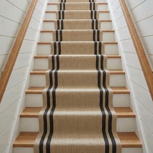 Puede incluir: Una alfombra para escaleras de material tejido en tono bronceado natural con dos franjas paralelas de color gris oscuro que recorren verticalmente el centro. La alfombra está instalada sobre peldaños de madera con contrahuellas blancas.