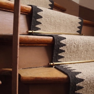 May include: A stair runner featuring a textured beige center with dark charcoal zig-zag patterned edges. The runner is secured to wooden stairs with polished brass rods that span the width of the carpet, creating a structured and functional appearance.