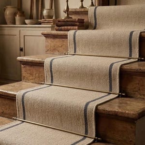 May include: A woven beige stair runner with two dark blue vertical stripes running down the center, secured to wooden stairs with horizontal brass rods. The runner features a textured, natural fiber appearance covering the treads and risers.