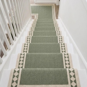 Puede incluir: Una alfombra de escalera color verde salvia con un borde color crema que presenta un patrón de diamantes repetitivo. La alfombra cubre el centro de los escalones de madera blanca, extendiéndose desde el rellano hasta el último escalón.