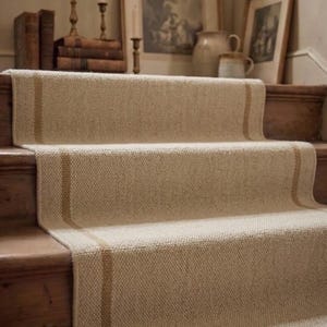 Könnte beinhalten: Ein gewebter Treppenläufer aus naturfarbenen beigen Fasern mit zwei vertikalen, hellbraunen Streifen, die über die gesamte Länge verlaufen. Der Läufer ist über Holzstufen verlegt und bedeckt die Tritt- und Setzstufen in einem durchgehenden Muster.