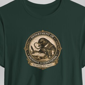 May include: A dark green t-shirt featuring a circular gold and black graphic of a crow looking through a magnifying glass at an open book. The text reads DEPARTMENT OF EXCESSIVE INTERPRETATION and NO DETAIL TOO SMALL in a decorative frame.
