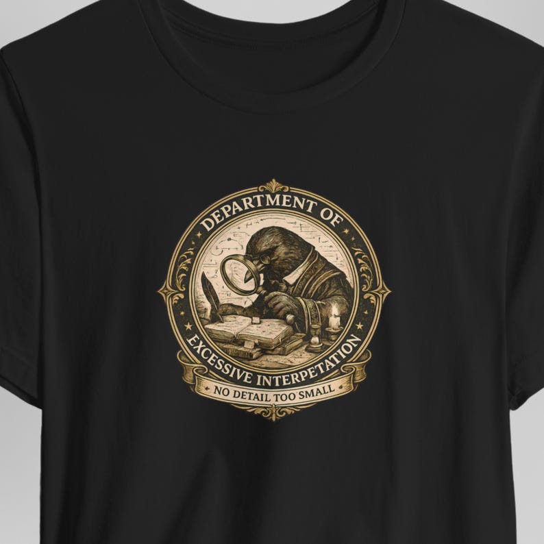 Close-up of a black graphic T-shirt featuring an ornate vintage gold and cream emblem with a scholarly crow studying an open book through a magnifying glass. The dark academia illustration includes the phrases “Department of Excessive Interpretation” and “No Detail Too Small.”