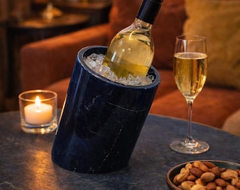 May include: A dark blue marble wine chiller with a bottle of white wine in ice. A lit candle, a glass of wine, and a bowl of nuts are on the table. The setting is a cozy, dimly lit room.