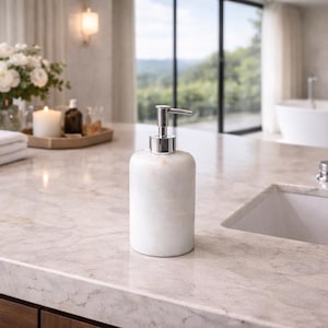 May include: A white marble soap dispenser with a silver pump. The dispenser is cylindrical and sits on a light-colored countertop. The background includes a sink, a window, and a tray with candles and flowers.