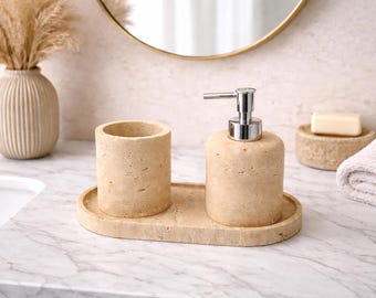 May include: A beige stone bathroom set includes a soap dispenser with a silver pump, a tumbler, and a tray. A white towel, soap, and a vase with dried flowers are also visible. The set is on a marble countertop.
