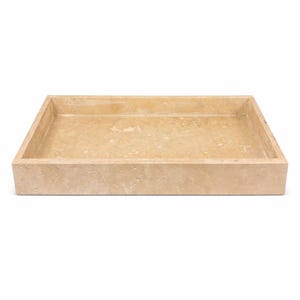 Pode incluir: Uma bandeja retangular de pedra bege com uma borda elevada. A bandeja tem uma superfície natural e texturizada, provavelmente feita de travertino. O design é simples e minimalista. Medidas em centímetros.