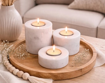May include: Three lit tea light candles in white marble holders of varying heights, arranged on a round wooden tray. A decorative vase with dried plants and a beaded garland with a tassel are also present.