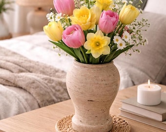 May include: A floral arrangement in a beige vase featuring pink and yellow tulips, white daisies, and a yellow daffodil. The vase sits on a woven coaster on a wooden table, with a lit candle and books nearby. The background includes a bed and a lamp.