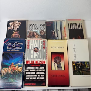 May include: A collection of eight music compact disc longbox cases featuring various artists including Willie Nelson, Carole King, Liberace, Bob James, and Rickie Lee Jones. The rectangular cases display album cover art, artist names, and song titles.