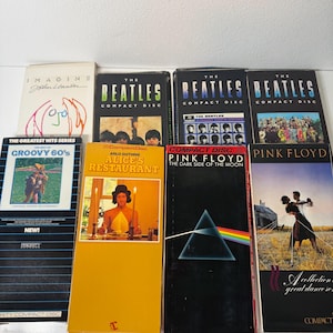 May include: A collection of eight music compact discs in long-box packaging. Titles include John Lennon's Imagine, three The Beatles albums, Groovy 60's, Arlo Guthrie's Alice's Restaurant, and two Pink Floyd albums, The Dark Side of the Moon and a collection.