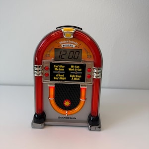 Puede incluir: Un despertador digital plateado y rojo con forma de gramola que muestra las 12:00. La unidad presenta el texto Musical Legends British Hits y los títulos de canciones Can't Buy Me Love, We Can Work It Out, A Hard Day's Night y Eight Days A Week.