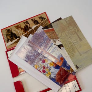 May include: A collection of rectangular greeting cards featuring various illustrations, including a group of kittens on a stone wall, a winter forest scene with trees, and a still life painting of yellow and red tulips in a blue bowl.