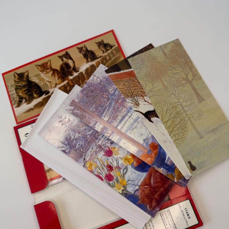 May include: A collection of rectangular greeting cards featuring various illustrations, including a group of kittens on a stone wall, a winter forest scene with trees, and a still life painting of yellow and red tulips in a blue bowl.
