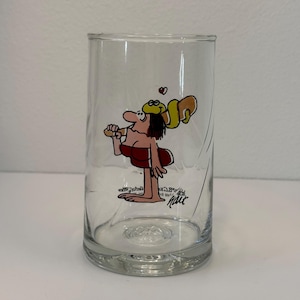 May include: A clear glass tumbler featuring a colorful cartoon graphic of a character with dark hair wearing a red top, carrying a yellow object on their shoulder with a small red heart above it, and the signature text "hart" printed below.