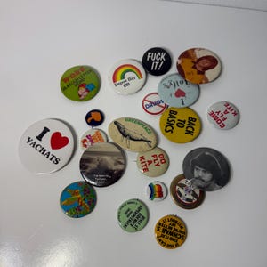 May include: A collection of round and oval pin-back buttons featuring various designs, including text such as I YACHATS, WORK IS A FOUR LETTER WORD, FUCK IT, GREENPEACE, BACK TO BASICS, GO FLY A KITE, and SCHWAB'S, alongside illustrations of a whale, rainbow, and people.