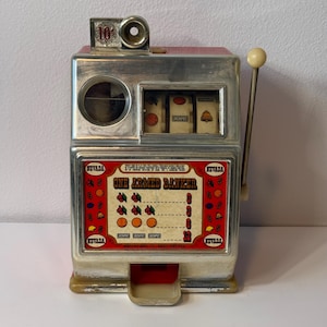 May include: A silver and red metal slot machine bank labeled One Armed Banker with a side pull lever, a coin slot marked 10 cents, and a front display showing cherry, orange, and bell symbols with payout values and the text Nevada Mfg Co Reno Nevada.