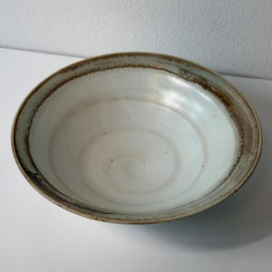 May include: A round ceramic bowl with a wide, shallow basin. The interior features a smooth, off-white glaze, while the wide, sloping rim is finished with a textured, dark brown reactive glaze that creates a distinct contrast against the center.