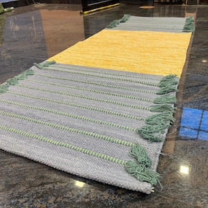 May include: A rectangular woven runner rug featuring a central block of bright yellow flanked by two larger sections of light gray. The gray areas are accented with horizontal rows of light green stitching and finished with matching green tassels.