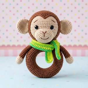 May include: A handmade crochet monkey rattle. The toy features a brown head and body with a tan face and ears. It has a green and yellow scarf and a ring-shaped body. The monkey has black safety eyes.