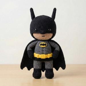 Puede incluir: Muñeco de Batman tejido a mano. El muñeco es principalmente negro y gris, con un cinturón amarillo y el logo de Batman en el pecho. Tiene una capa negra y una capucha con orejas puntiagudas. El muñeco está sobre una superficie marrón claro.