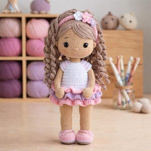 May include: A handmade crochet doll with brown curly hair, wearing a white dress with pink and purple ruffles, and pink shoes. The doll has a headband with a flower. The background includes yarn and crochet hooks.