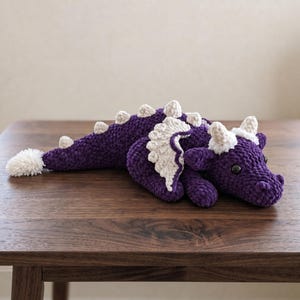Pode incluir: Um brinquedo de pelúcia de dragão de crochê roxo feito à mão com detalhes brancos. O dragão tem um corpo longo com espinhos brancos, asas e uma cauda fofa. O dragão está sobre uma superfície de madeira escura.