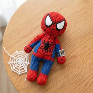 May include: A crocheted Spider-Man doll featuring a red and blue yarn body with a black spider emblem on the chest, white yarn eyes, and a separate white crocheted spider web. A small fabric tag on the arm reads DAILY BUGLE PRESS.