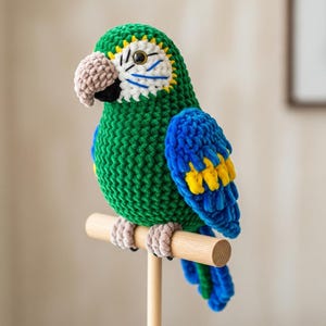 May include: A handmade, green, blue, and yellow crochet parrot perched on a wooden dowel. The parrot has a tan beak, white and blue accents around the eyes, and tan feet. The wings are blue with yellow details.