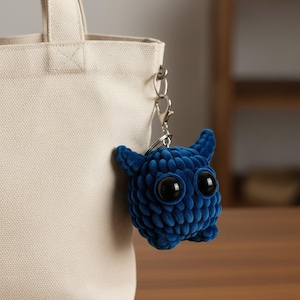 Puede incluir: Un llavero de monstruo azul tejido a ganchillo con grandes ojos negros y pequeños cuernos, sujeto a un cierre plateado. El peluche cuelga de un bolso tote beige, con un fondo de madera.