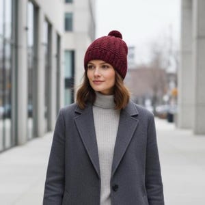 May include: A woman wearing a burgundy knit beanie with a pom-pom, a gray turtleneck sweater, and a dark gray coat. The beanie has a textured pattern. The background shows a city street with buildings.