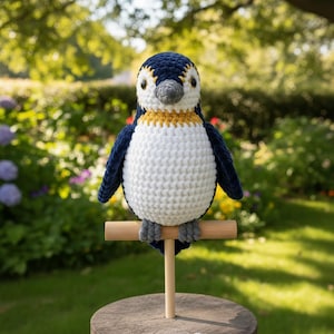 May include: A handmade crochet penguin with a white belly, navy blue wings and back, and a yellow and white head. The penguin is perched on a wooden dowel, mounted on a wooden base, set against a blurred garden background.