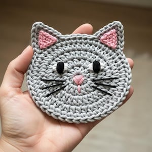 May include: Handmade gray crochet cat face with pink ears, black eyes, and whiskers. The cat face is round and has a pink nose. The crochet stitches are visible, creating a textured appearance. This item is a coaster or decorative piece.
