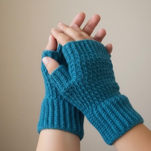Könnte beinhalten: Ein Paar türkisfarbene Fäustlinge. Die Handschuhe sind mit einem gerippten Bündchen und einem strukturierten Stich auf der Hand gestrickt. Die Handschuhe sind so konzipiert, dass sie die Hände warm halten und gleichzeitig die Finger frei lassen.