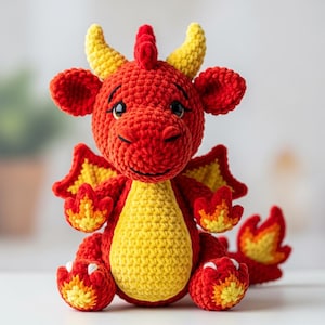 Könnte beinhalten: Ein handgehäkelter Drache in leuchtendem Rot und Gelb. Das Plüschtier hat detaillierte Flügel, Hörner und einen freundlichen Gesichtsausdruck. Der Drache sitzt und zeigt seinen flammenartigen Schwanz und seine Füße. Eine charmante, skurrile Kreation.