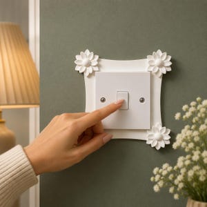 Puede incluir: Un interruptor de luz blanco con un marco decorativo blanco adornado con cuatro acentos florales. Un dedo presiona el interruptor en una pared verde. El interruptor está centrado dentro del marco, con flores en cada esquina.