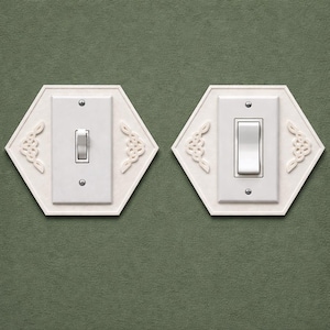 May include: Two white light switch covers with a decorative hexagonal design. Each cover has a rectangular switch plate with a toggle switch. The covers are mounted on a green wall.
