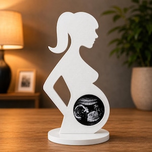 Può includere: Silhouette bianca di una persona incinta, con una coda di cavallo e un'apertura rotonda che mostra un'immagine ecografica. La figura è montata su una base circolare, un oggetto decorativo.