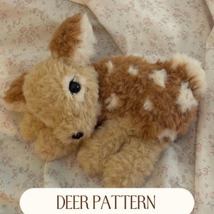 Pode incluir: Um peluche em forma de cervo com pelo macio bege e castanho, com manchas brancas nas costas, olhos escuros e um pequeno nariz preto. O texto DEER PATTERN é exibido num oval branco na parte inferior da imagem.