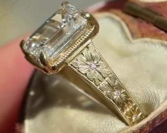 Pode incluir: Um anel de ouro com uma grande pedra preciosa transparente retangular de corte esmeralda num engaste de garras. A banda é feita de ouro amarelo e apresenta um padrão floral gravado intrincado na superfície, acentuado com pequenas pedras incrustadas.