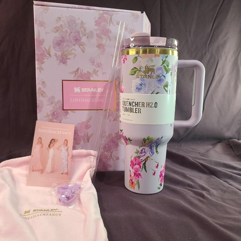 May include: A lavender Stanley Quencher H2.0 tumbler with a floral pattern, a handle, and a clear straw. It is accompanied by a matching floral gift box, a small informational card featuring three figures, a light pink fabric pouch, and a small purple accessory.