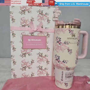 May include: A cream-colored Stanley Quencher tumbler with a pink floral and bow pattern, featuring a matching handle, lid, and straw. The tumbler is displayed next to a tall, rectangular gift box with the same floral print and a pink ribbon.