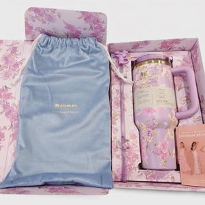May include: A lavender Stanley tumbler with a floral pattern, a matching straw, and a light blue drawstring pouch inside a floral gift box. The box includes a card featuring three people in dresses and the text LoveShackFancy and Stanley.